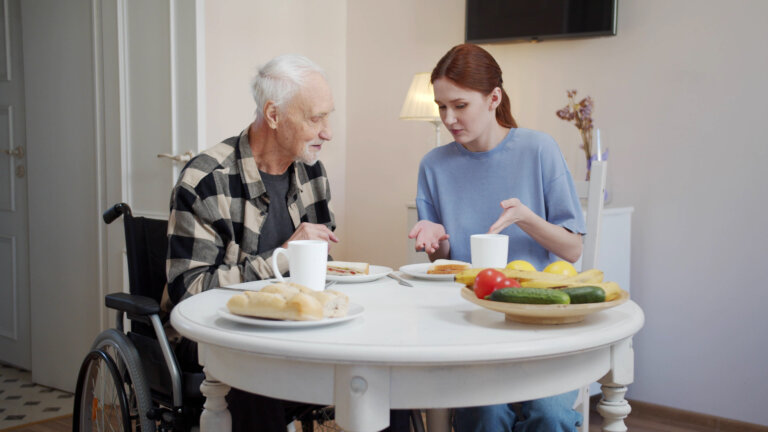 A woman enters the dining room and puts plates of sandwiches on the table. She sits down and starts talking to an older man with glasses. They discuss food. High quality 4k footage